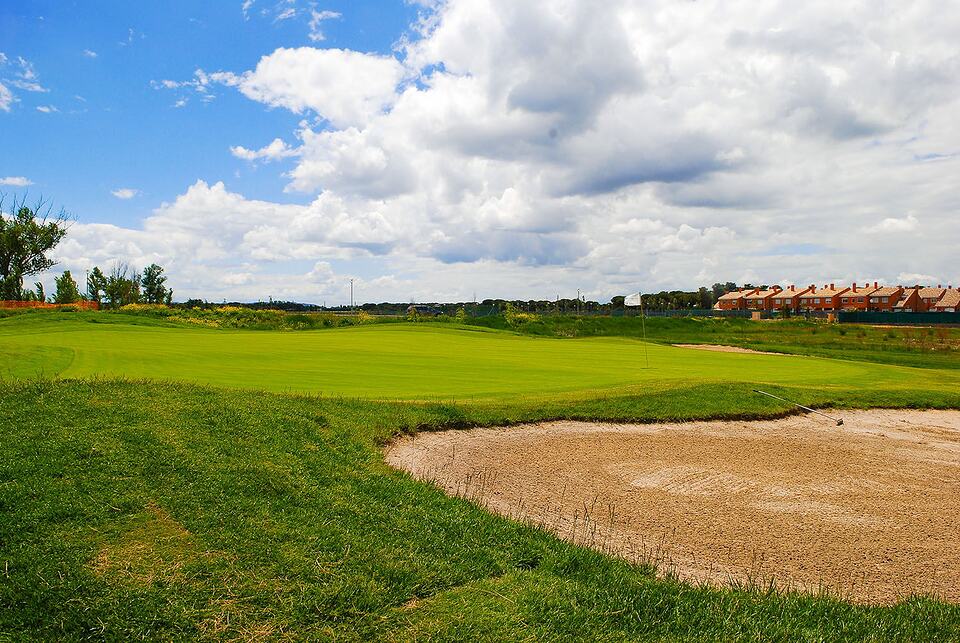 A golf green beside a sand bunker, with houses and a cloudy blue sky in the background.
