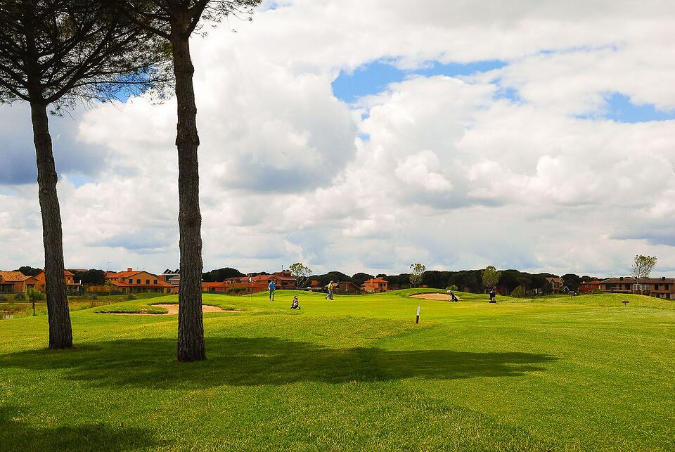 Aldeamayor golf course with golfers crossing the fairway beneath tall trees and a cloudy sky