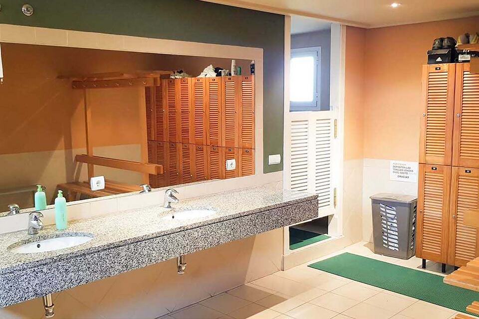 Club changing room with wooden lockers, a bench and a long double sink counter.