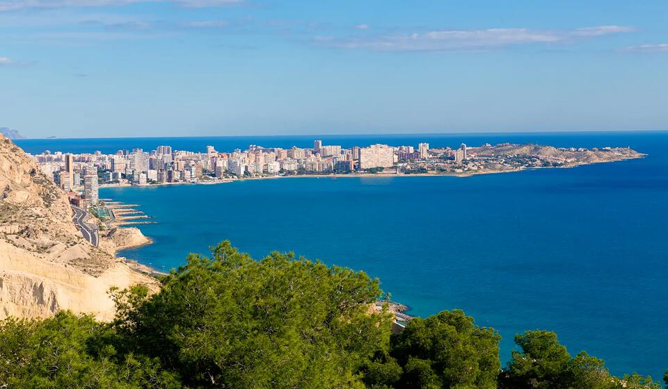Coastal city seen from above, with a rocky headland, blue sea and a long stretch of high-rise buildings.