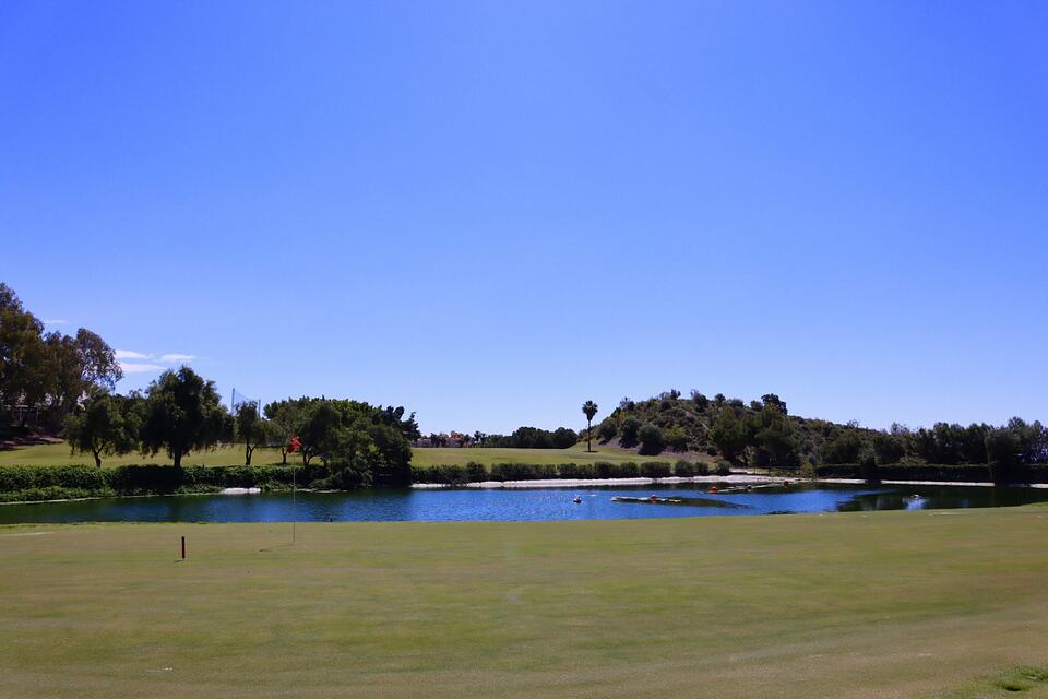 A golf course beside a pond, with trees and a low hill in the background.