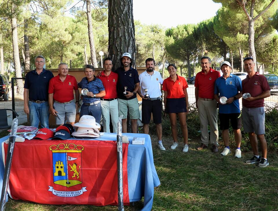Golfers pose with trophies beside a table of prizes under pine trees.