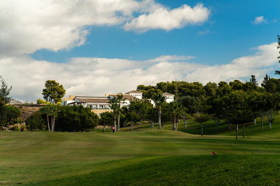 Baviera Golf clubhouse beyond a grassy golf course with palm trees and wooded hills