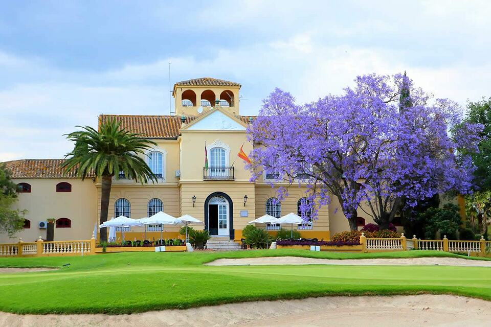Clubhouse of Real Guadalhorce Golf Club beside a green, with a purple-flowering tree and sand bunker
