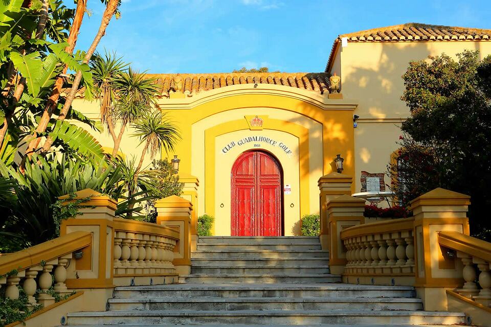Yellow clubhouse entrance with a red wooden door, tiled roof and stone steps.