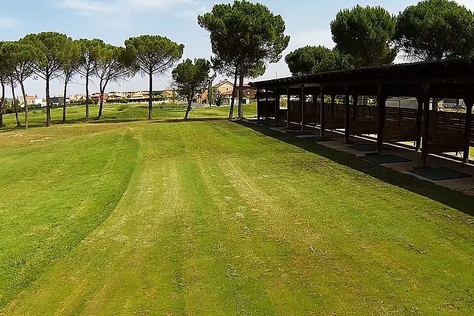Covered bays at the golf driving range beside a grassy practice area and pine trees.