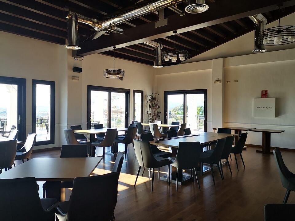 Dining room at Baviera Golf with tables, chairs and large windows opening onto a terrace.