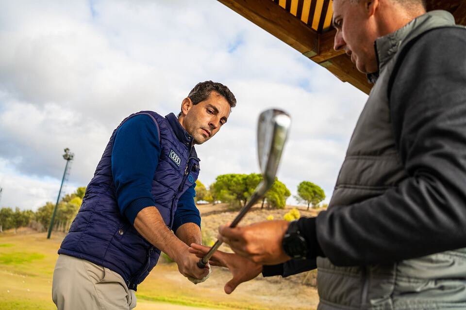 A golf instructor guides an adult player as he holds a club at the practice range.