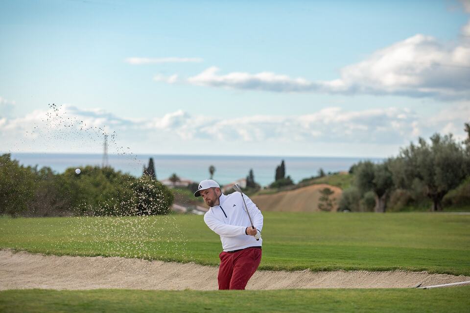 Golfer splashing sand from a bunker with the sea visible in the background