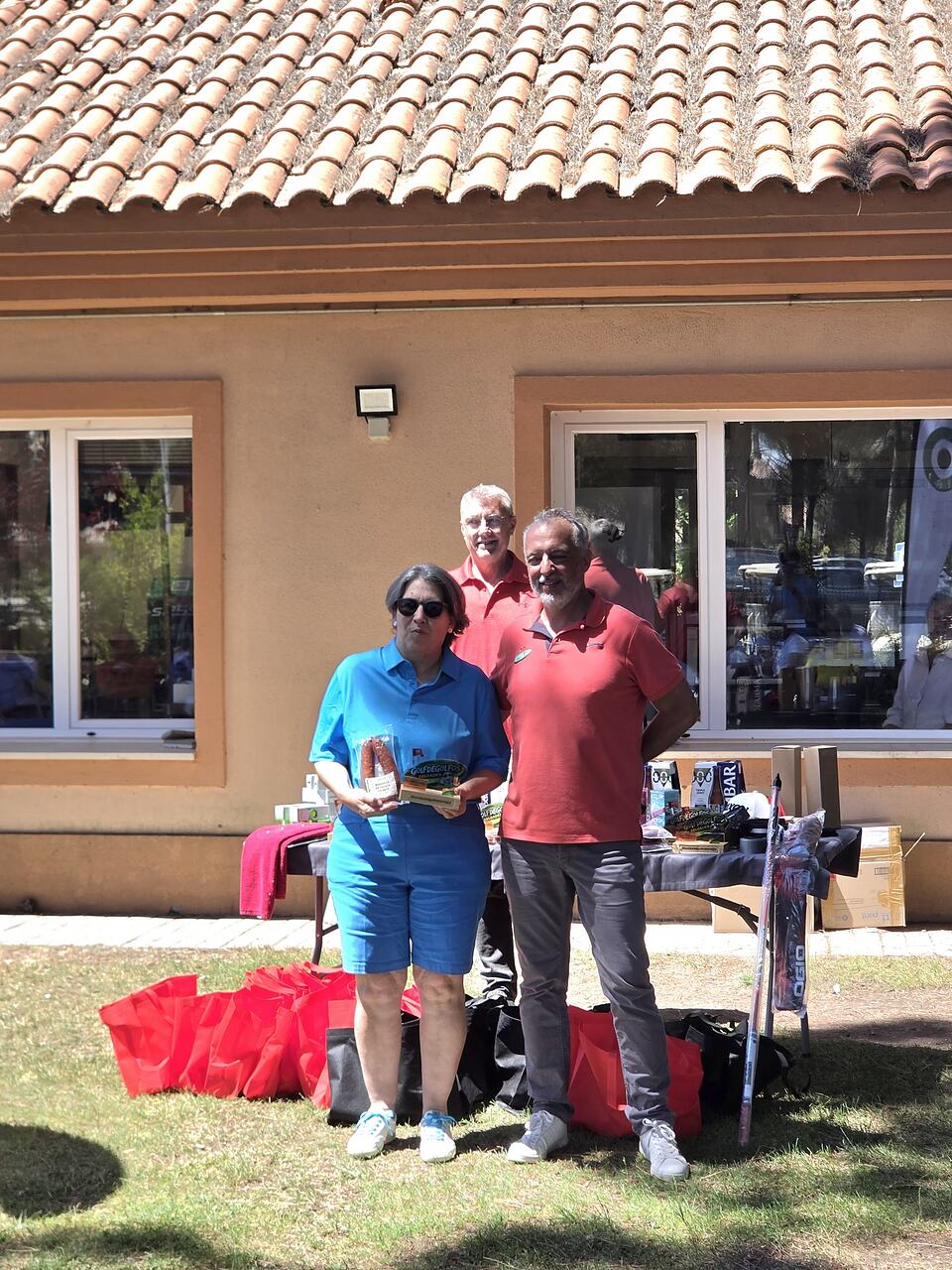 Three men pose with prizes in front of the clubhouse after a golf event.