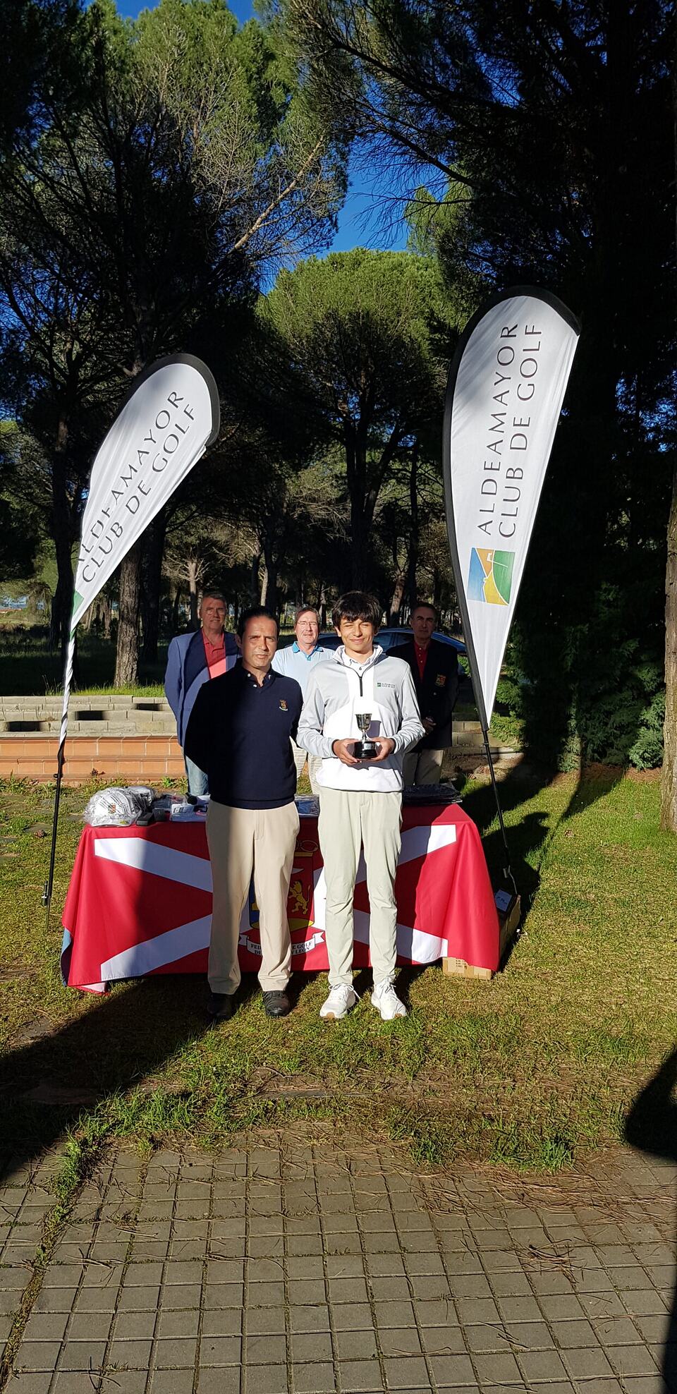 A golf tournament winner holds a trophy with club staff beside Aldeamayor Golf Club banners.