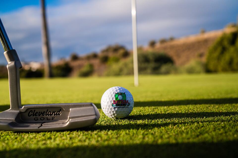 Golf ball and Cleveland putter on the green at Baviera Golf, with a flag and hills in the background.