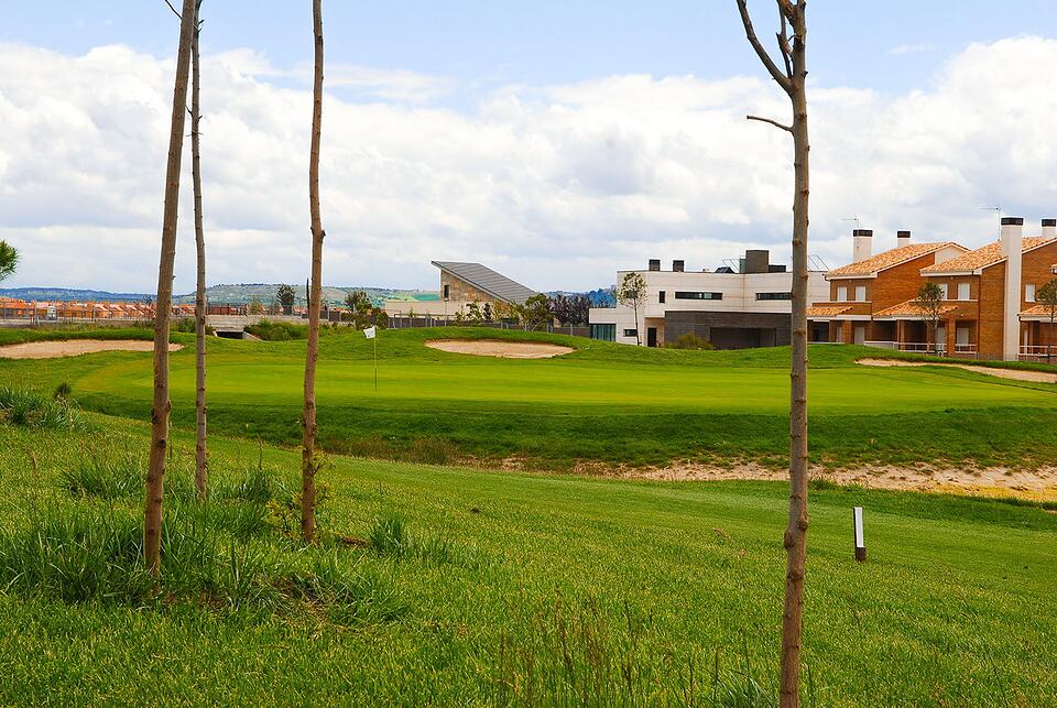 A green with a flag and sand bunkers beside modern buildings at Aldeamayor Golf Club.