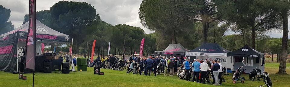Golf event with players gathered among sponsor tents and golf bags on a lawn beneath pine trees.