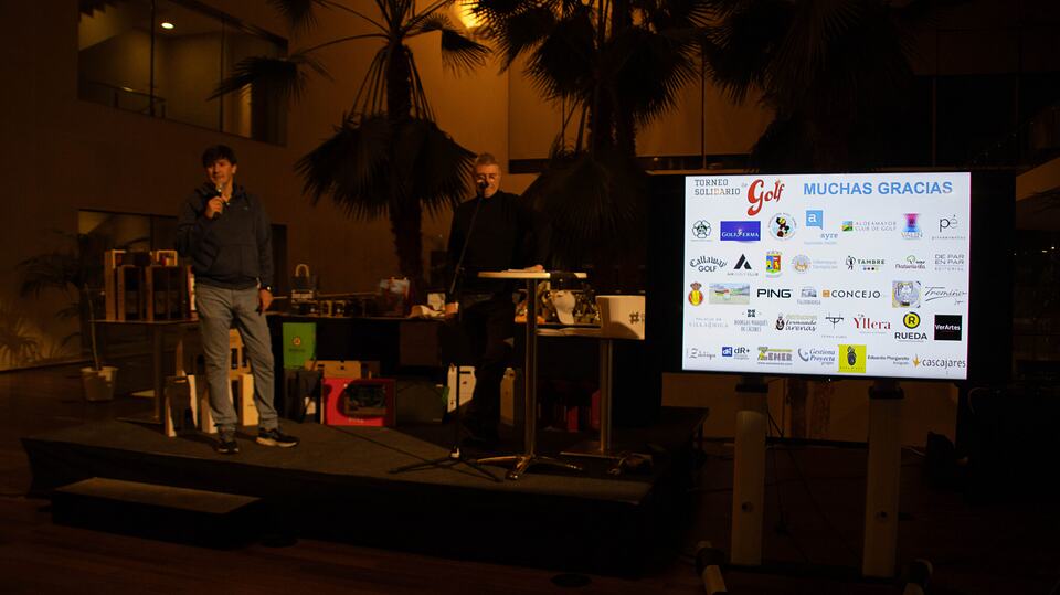 Dos hombres en un escenario del torneo solidario de Aldeamayor Club de Golf, junto a una pantalla con logotipos
