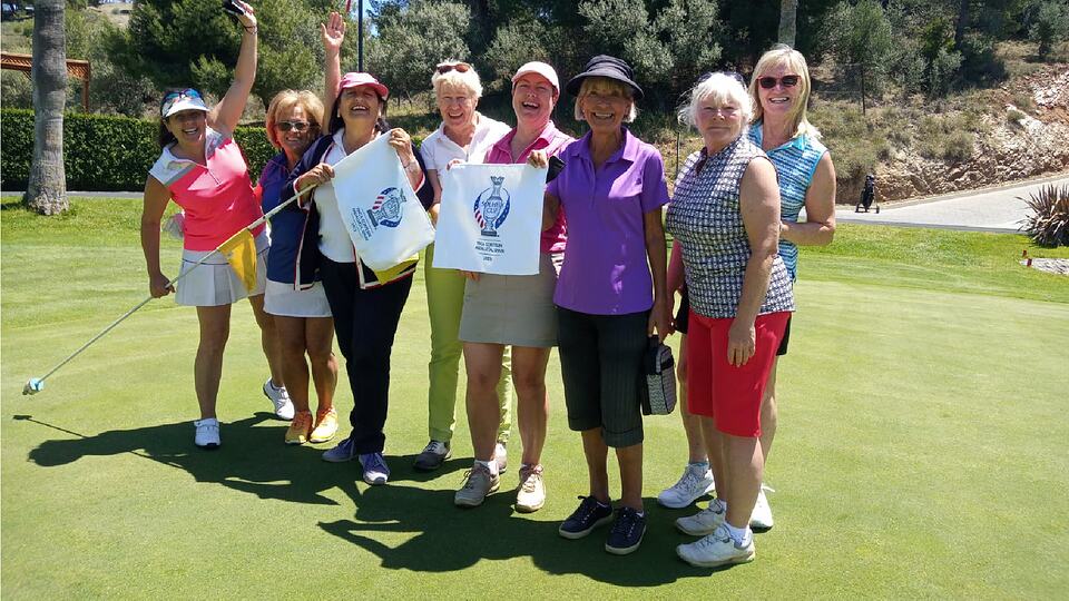 A group of women golfers pose with golf flags and prize bags on a sunny green.