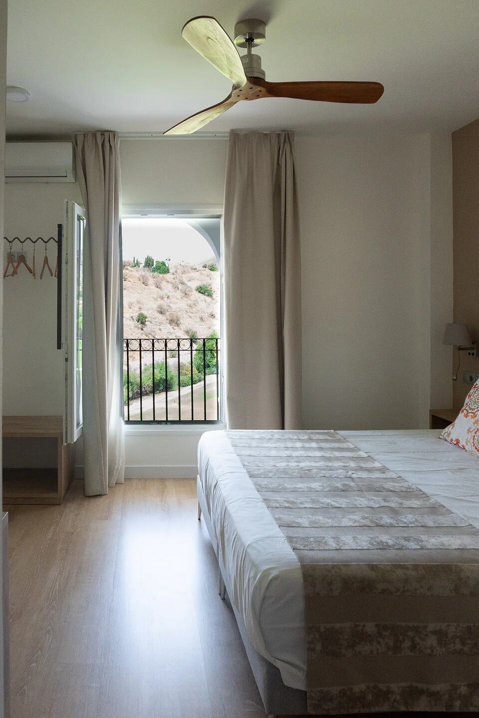 Bright minimalist bedroom with a double bed and an open balcony overlooking a dry hillside