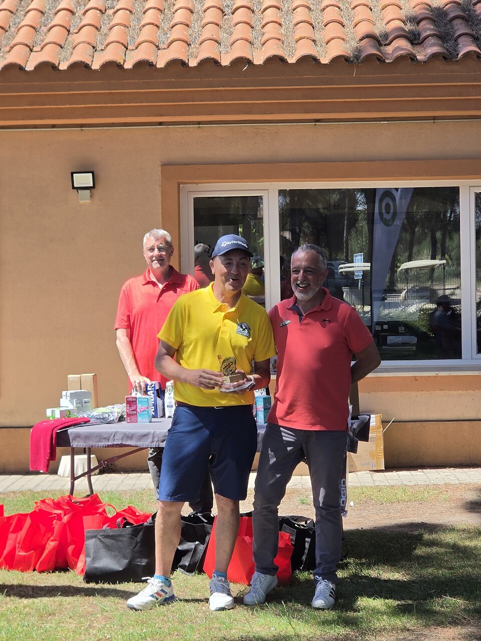 Three men pose with a trophy at a golf event outside the clubhouse.
