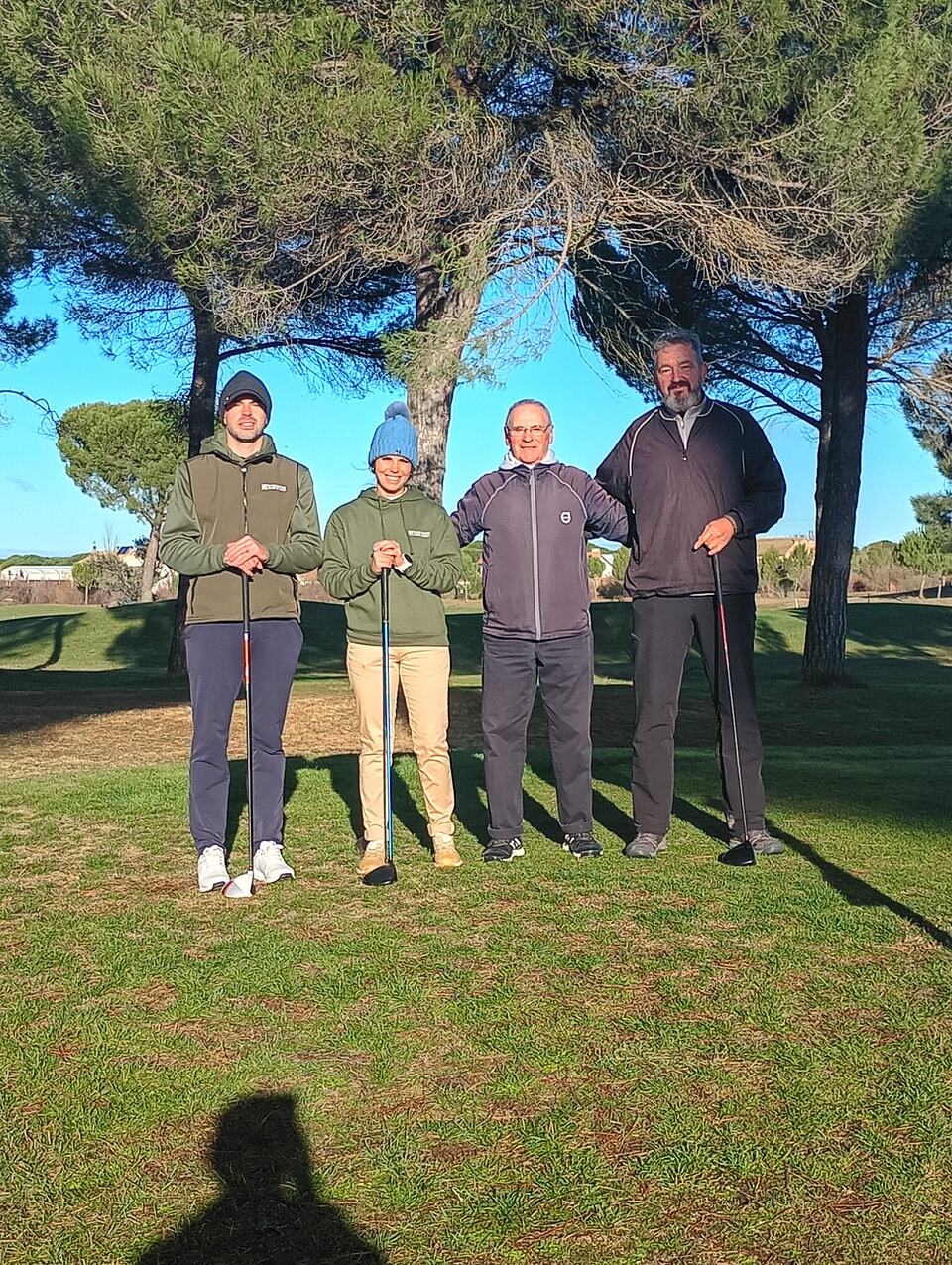Four golfers pose with clubs beneath pine trees on the course.