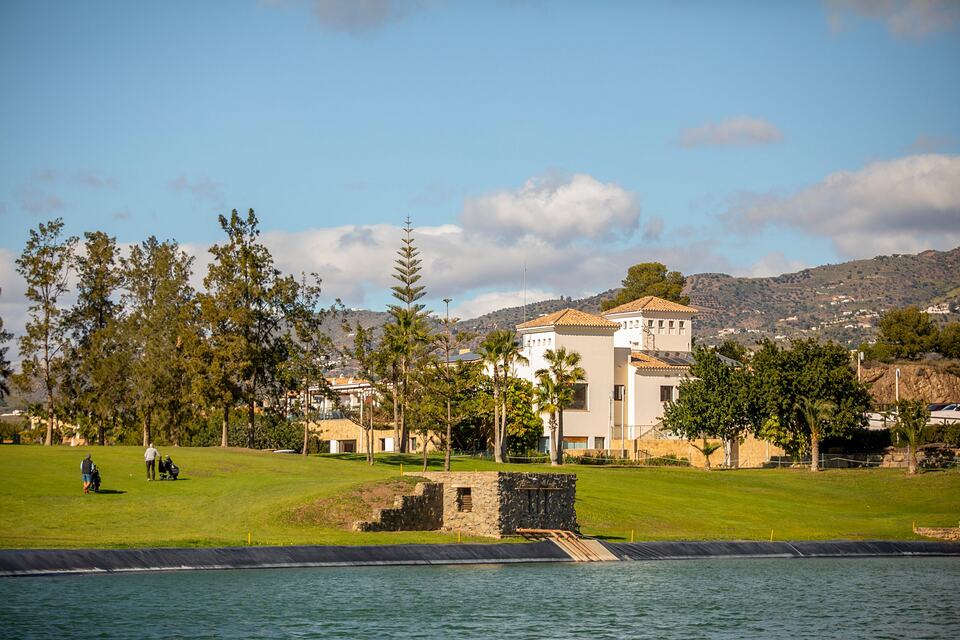Golfers on a green beside a water hazard, with a clubhouse and hills in the background.