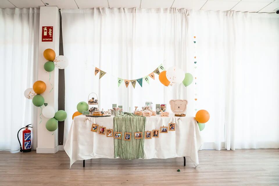 A decorated event table with balloons, a bunting banner and party favors in a bright hall.
