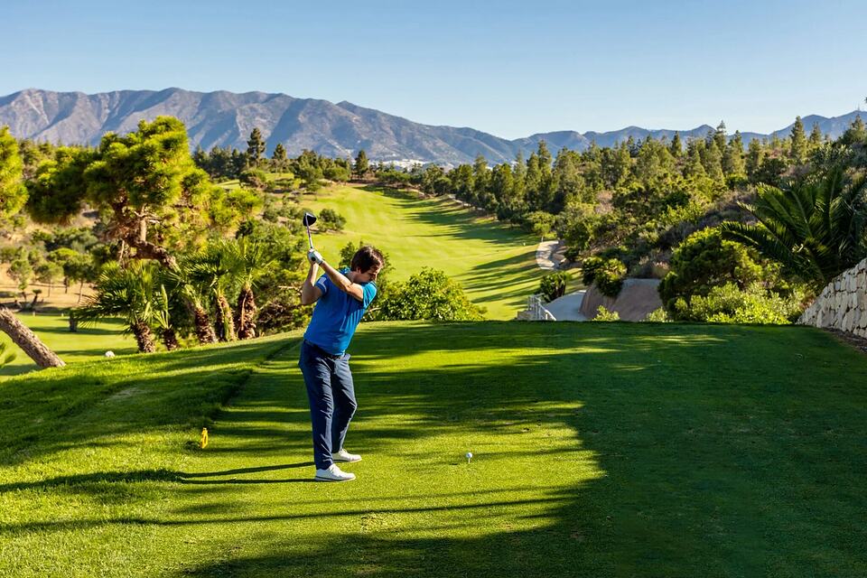 Golfer swinging from a tee on a green course with mountains in the background