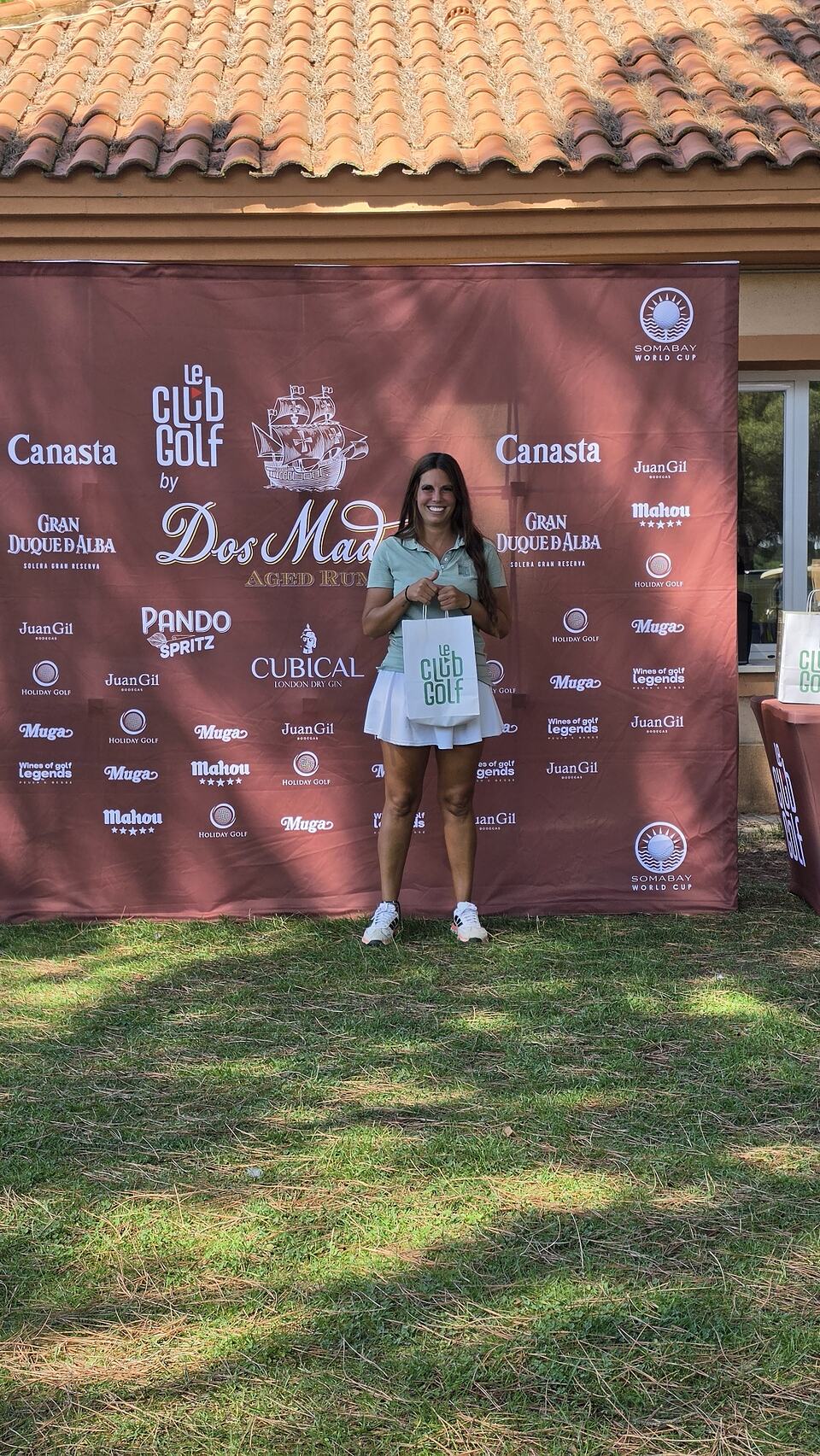 Mujer con una bolsa de Le Club Golf frente a un panel de patrocinadores del torneo