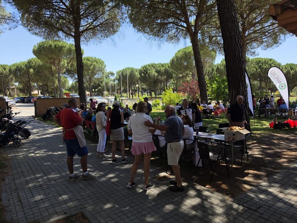 Golfers gather beneath pine trees at an outdoor tournament event.