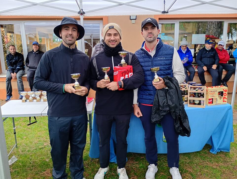 Three golf tournament winners pose with trophies beside a prize table, with spectators behind them.