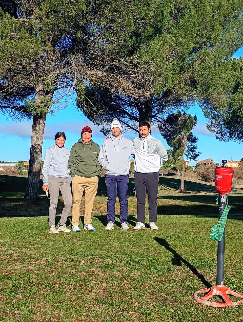 Four Ryder event participants pose together on the golf course beneath pine trees.