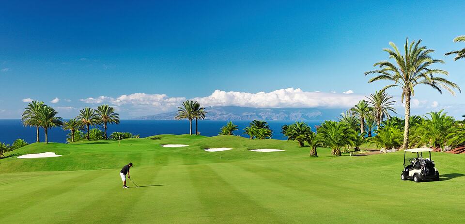 Un golfista en un green con vistas al mar, palmeras, bunkers y un buggy.