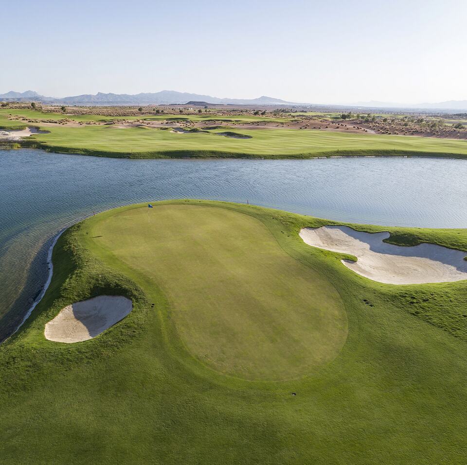 Aerial view of a golf green beside a lake, with bunkers and mountains in the distance at Alhama Signature Golf.