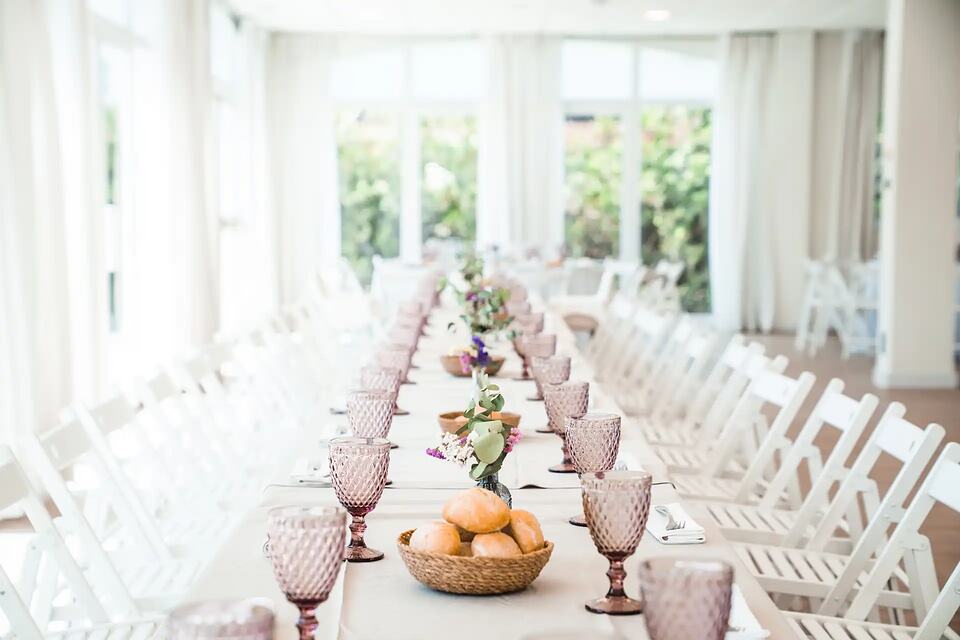 Long banquet table set with pink glasses, flowers and bread in a bright event room